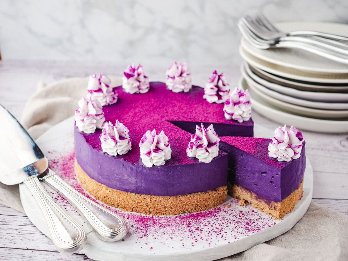 Sliced ube cheesecake on a serving board, with utensile on the side and a stack of plates and forks in the background.
