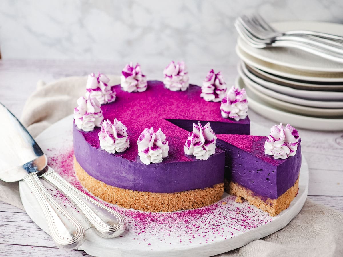 No Bake Cheesecake Recipes 27 Sliced ube cheesecake on a serving board, with utensile on the side and a stack of plates and forks in the background.