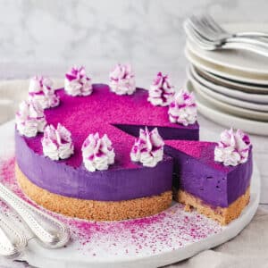 Sliced ube cheesecake on a serving board, with utensile on the side and a stack of plates and forks in the background.