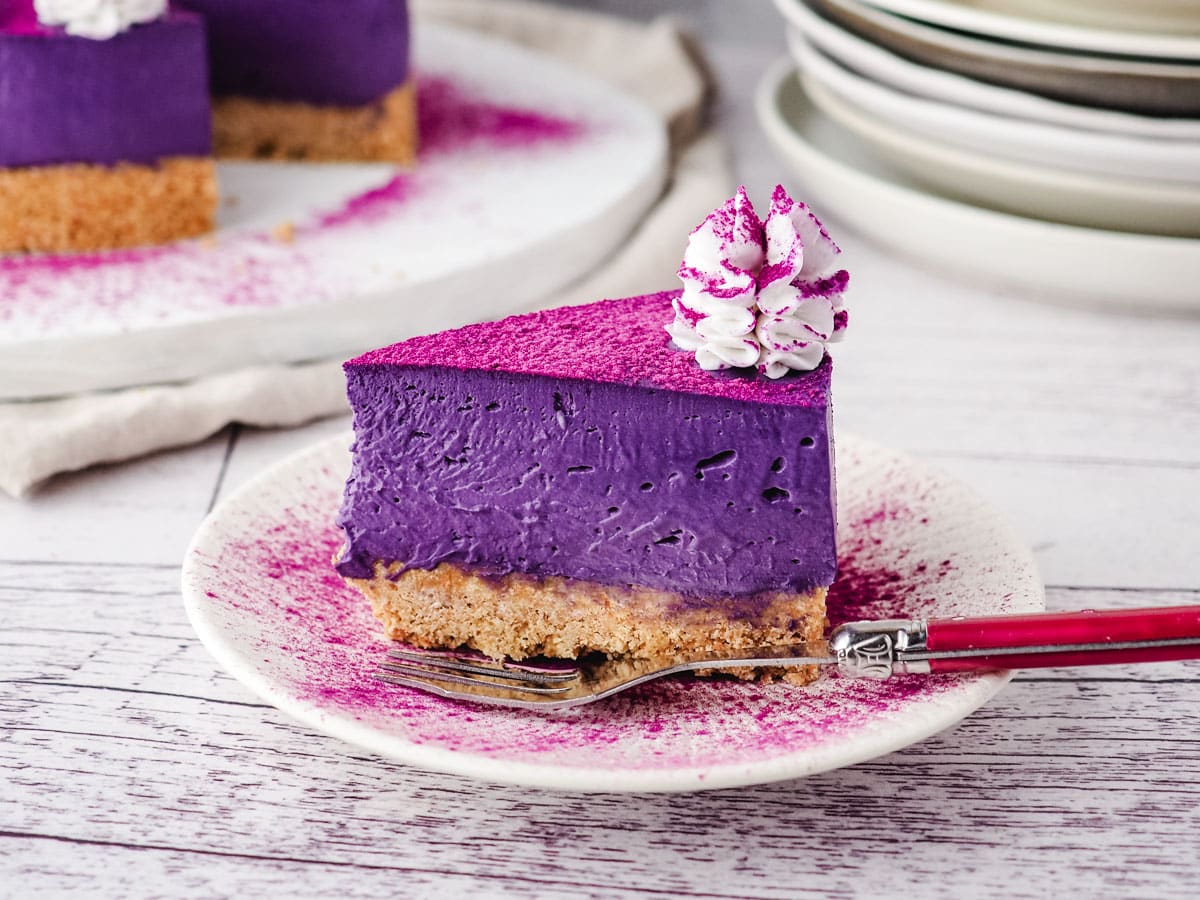 Purple yam cheesecake on plate with a fork on the side and rest of back and a stack of plates in the background.