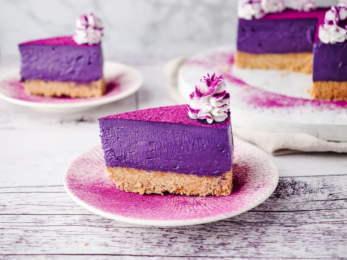 Slice of ube cheesecake on a plate dusted with ube powder, with rest of cake and another slice of cake in the background.