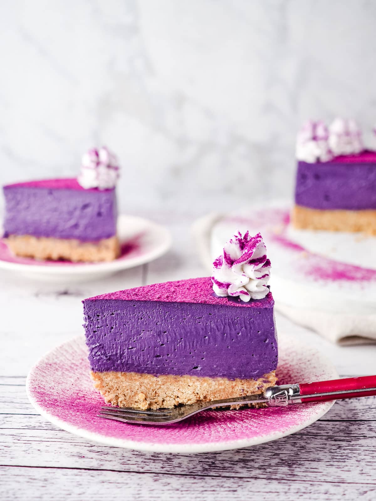 Slice of ube cheesecake on a plate dusted with ube powder, with a fork on the sideand rest of cake and another slice of cake in the background.