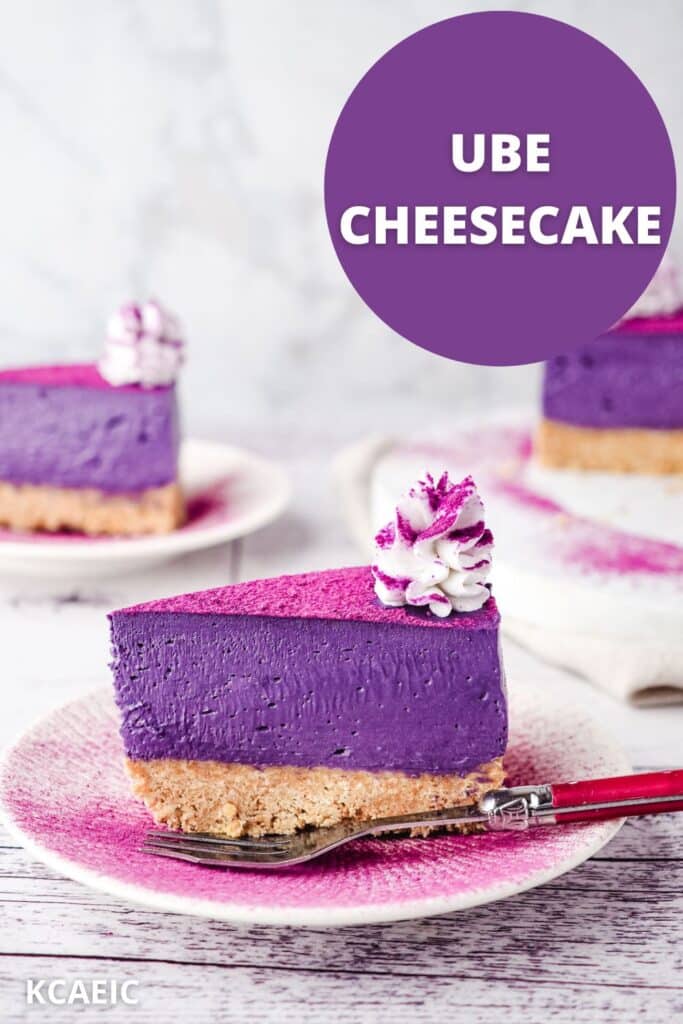 Slice of ube cheesecake on a plate dusted with ube powder, with a fork on the sideand rest of cake and another slice of cake in the background.