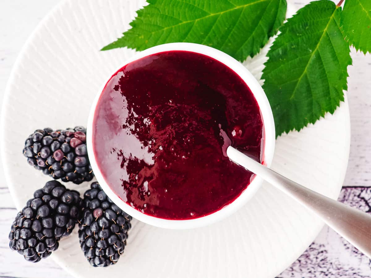 Blackberry topping for ice cream in a serving bowl with a spoon and fresh blackberries and leaves on the side.