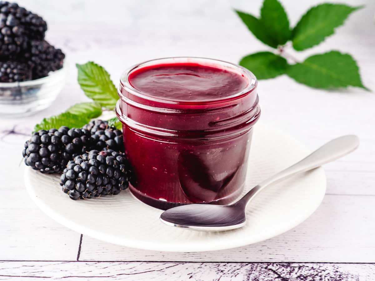 A small mason jar full of blackberry coulis, with a spoon and fresh blackberries on the side.