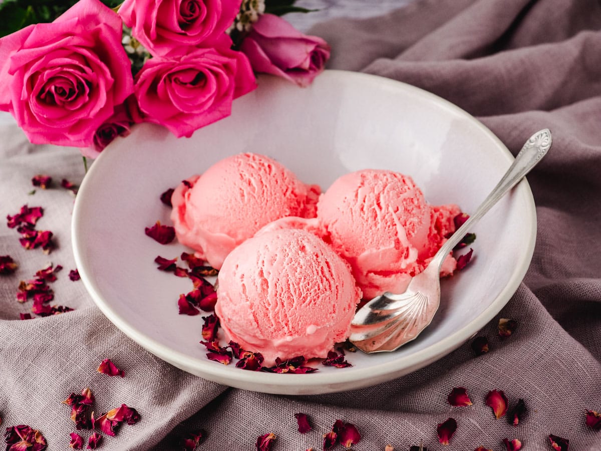 Scoops of rose ice cream in a bowl and a spoon on the side, garnished with rose petals and fresh roses in the background.