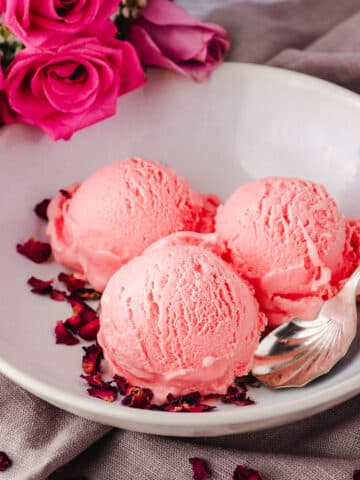 Scoops of rose ice cream in a bowl and a spoon on the side, garnished with rose petals and fresh roses in the background.