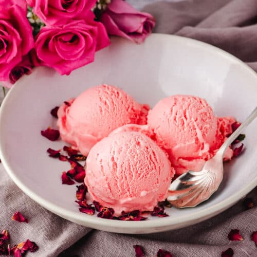 Scoops of rose ice cream in a bowl and a spoon on the side, garnished with rose petals and fresh roses in the background.