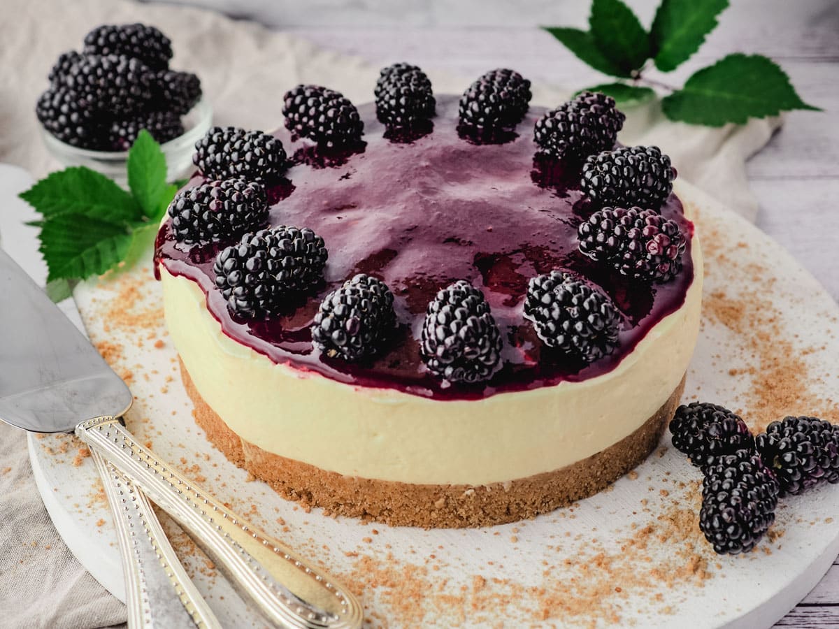 Whole no bake blackberry cheesecake with blackberry coulis and fresh blackberries on a serving plate, with serving ware on the side.