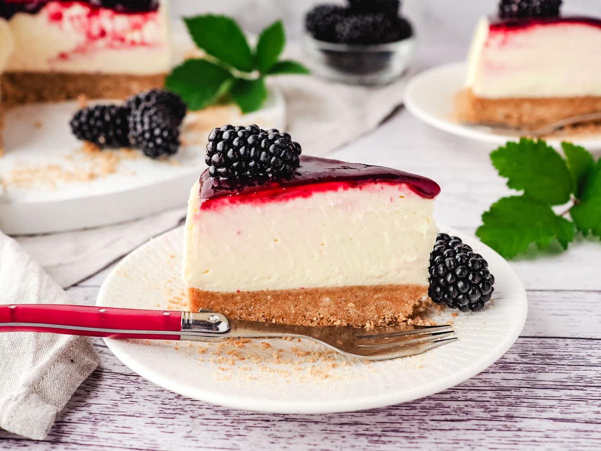 Slice of no bake blackberry cheesecake on a plate with fork, and rest of cake in background.