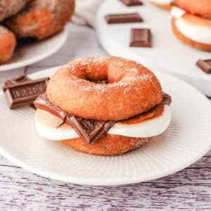 Smore Donuts 10 A smore donut on a plate with pieces of Hershey’s chocolate on the side and more smores donuts on a plate in the background.