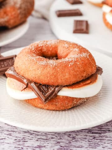 A smore donut on a plate with pieces of Hershey’s chocolate on the side and more smores donuts on a plate in the background.