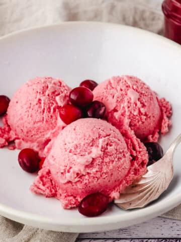 Scoops of cranberry ice cream in a bowl garnished with whole cranberries, with cranberry sauce and sugared cranberries on the side.