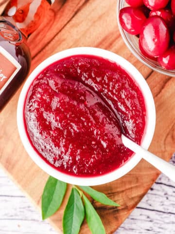 Homemade cranberry sauce with Grand Marnier in a serving dish and a spoon, with whole cranberries and bottle of Grand Marnier on the side.