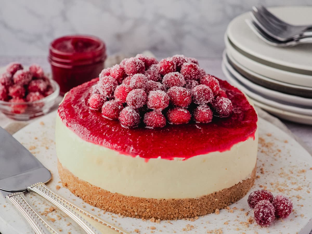 Sugared Cranberries 7 Cheesecake decorated with sugared cranberries and cranberry sauce, with a pile of plates, bowl of sugared cranberries and cranberry sauce in the background.