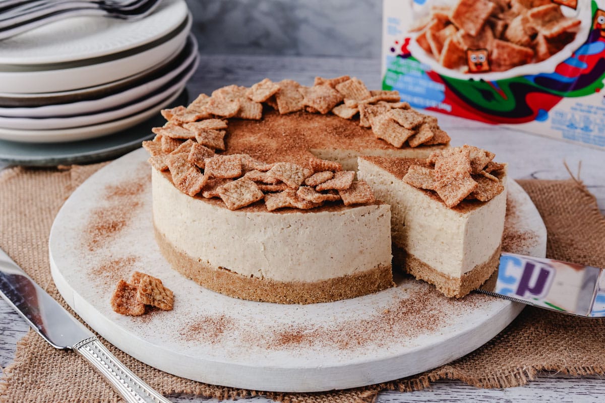 No Bake Cheesecake Recipes 6 Serving a slice of Cinnamon Toast Crunch cheesecake from a serving board, with a pile of plates and box of Cinnamon Toast Crunch in the background.