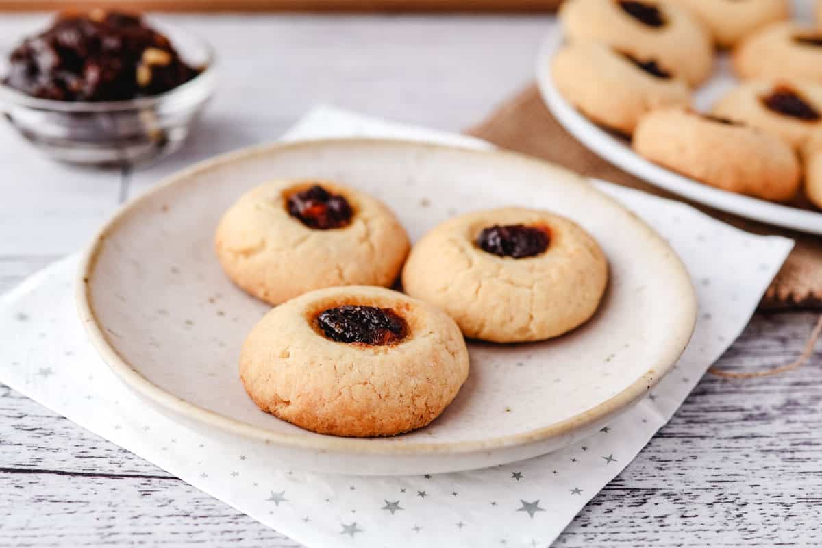 Mincemeat Thumbprint Cookies 2 Three mincemeat thumbprints on a plate with bowl of fruit mince and rest of cookies in the background.