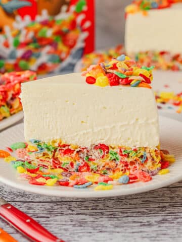 Slice of Fruity Pebbles cheesecake on a plate, with rest of cake and Rice Krispie Treats with Fruity Pebbles in the background.