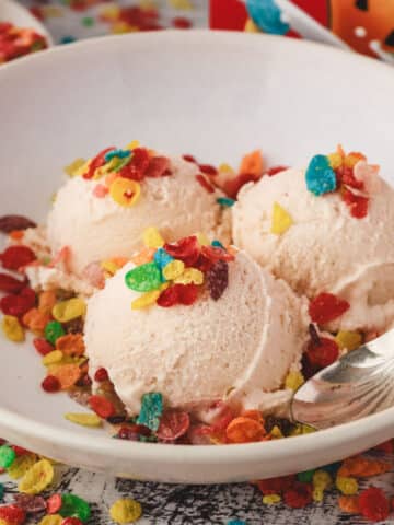 Three scoops of Fruity Pebbles ice cream decorated with Fruity Pebbles cereal, in a bowl with a spoon on the side.