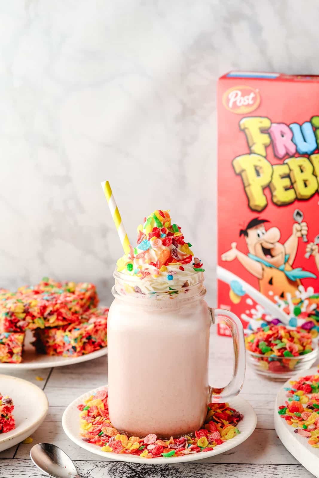 Fruity Pebbles Milkshake 8 Fruity Pebbles milkshake in a glass with a striped straw, decorated with whipped cream and Fruity Pebbles, with a box of Fruity Pebbles and other Fruity Pebbles desserts in the background.