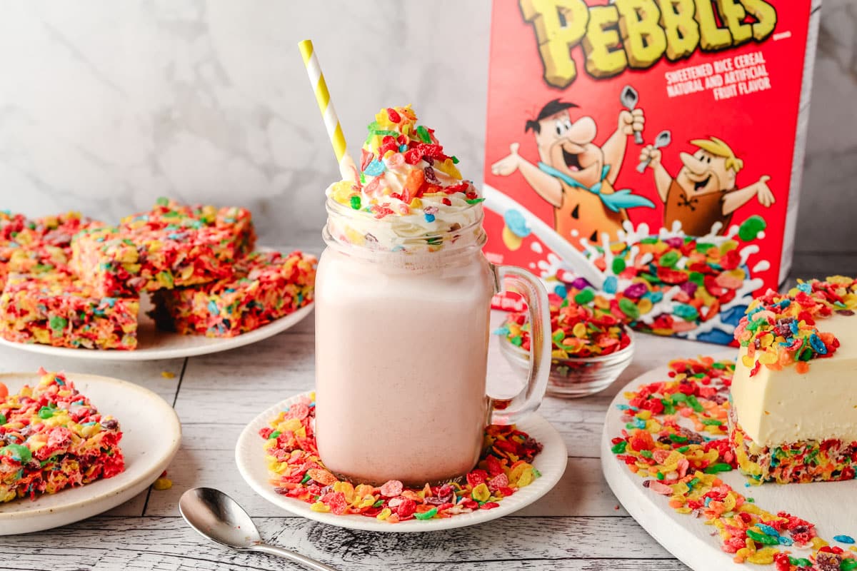 Fruity Pebbles Milkshake 2 Fruity Pebbles milkshake in a glass with a striped straw, decorated with whipped cream and Fruity Pebbles, with a box of Fruity Pebbles and other Fruity Pebbles desserts in the background.