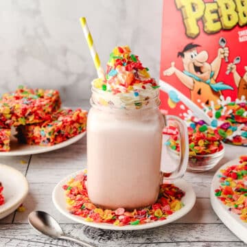Fruity Pebbles Milkshake 7 Fruity Pebbles milkshake in a glass with a striped straw, decorated with whipped cream and Fruity Pebbles, with a box of Fruity Pebbles and other Fruity Pebbles desserts in the background.