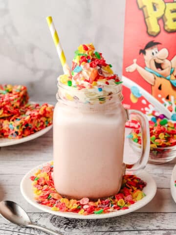 Fruity Pebbles milkshake in a glass with a striped straw, decorated with whipped cream and Fruity Pebbles, with a box of Fruity Pebbles and other Fruity Pebbles desserts in the background.
