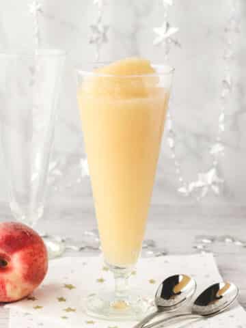 Glass of Bellini slushy with tall spoons and fresh peaches on the side.
