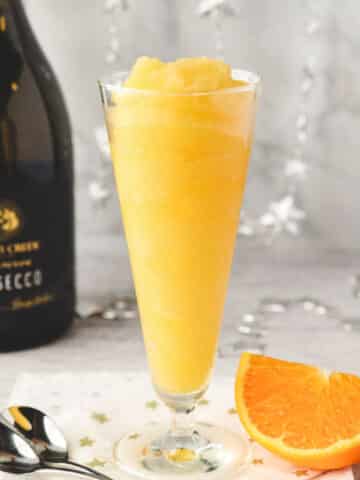 Glass of Mimosa slushy with tall spoons and fresh orange slice on the side.