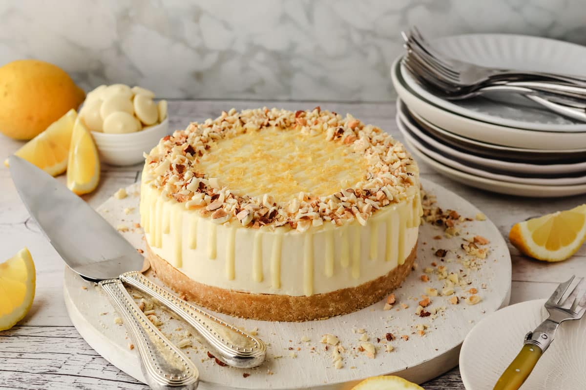 Lemon and White Chocolate Cheesecake 2 A lemon and white chocolate cheesecake with chopped lemon white chocolate almonds on top sits on a white board, surrounded by lemon slices, cutlery, and stacked plates.