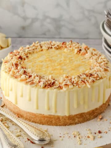 A lemon and white chocolate cheesecake with chopped lemon white chocolate almonds on top sits on a white board, surrounded by lemon slices, cutlery, and stacked plates.