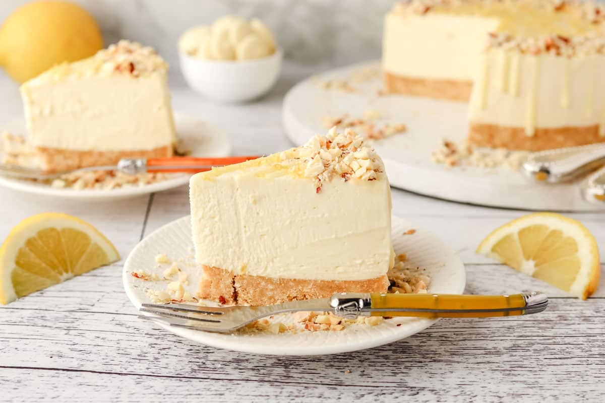 Lemon and White Chocolate Cheesecake 8 A slice of lemon white chocolate cheesecake on a plate, with rest of the cake in the background.