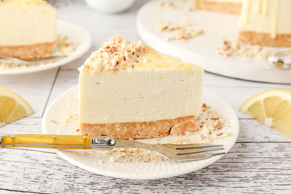 Lemon and White Chocolate Cheesecake 7 A slice of lemon white chocolate cheesecake on a plate, with rest of the cake in the background.