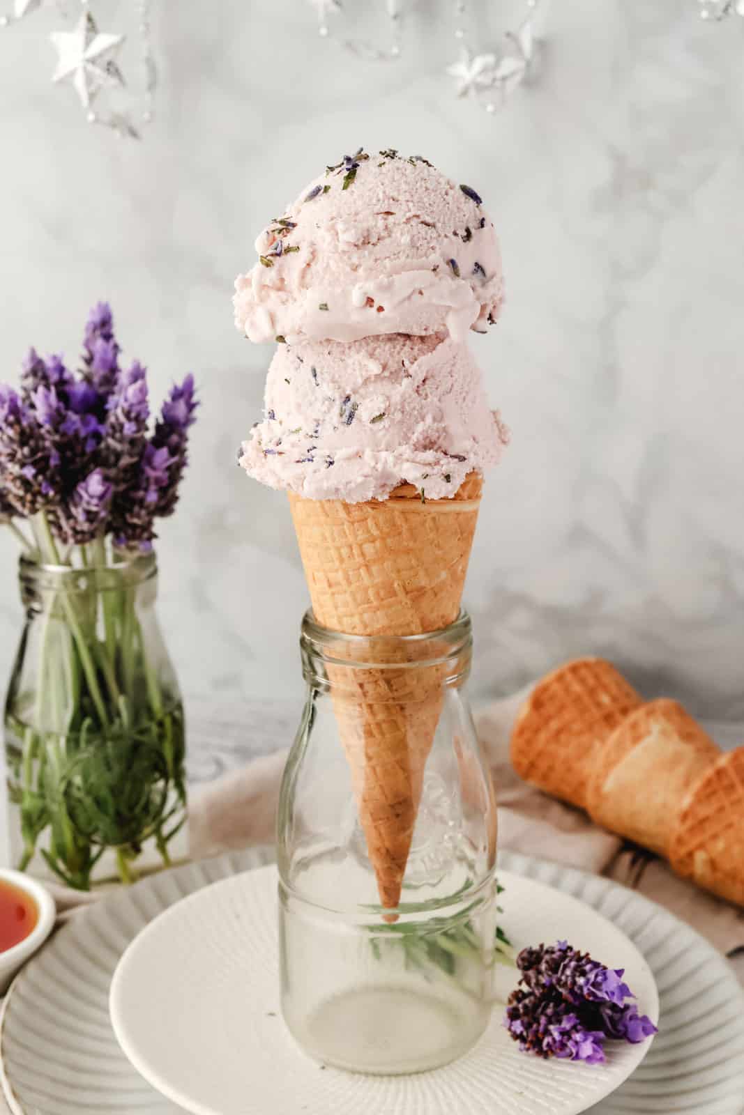 2 scoops of lavender ice cream in a cone with fresh lavender flowers in the background.