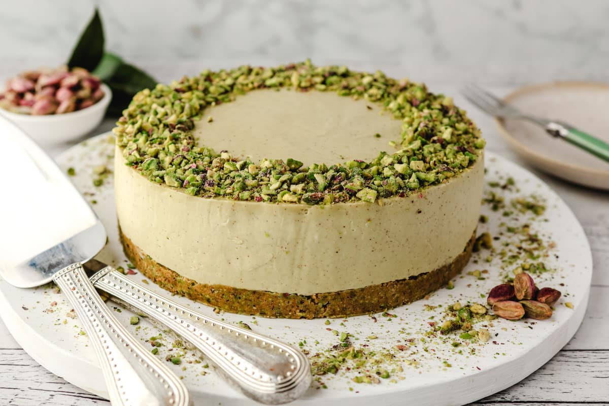 Whole no bake pistachio cheesecake on a serving board, with serving wear on the side.