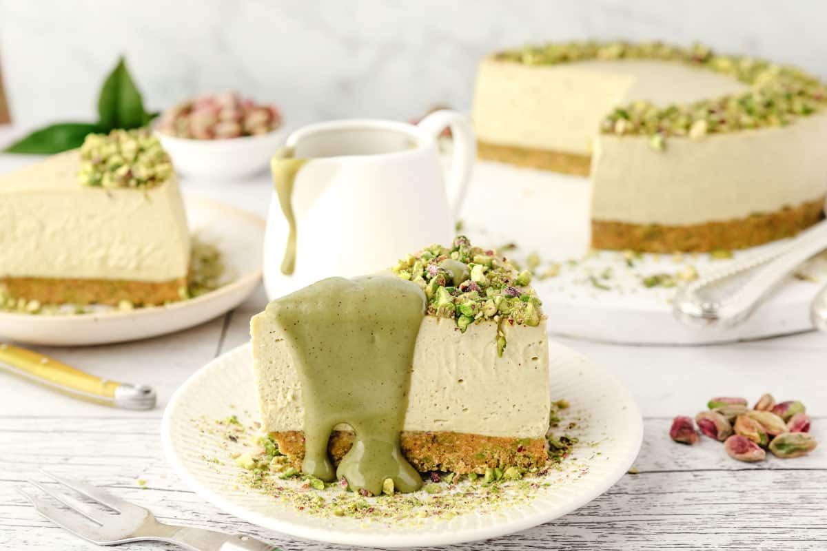 Slice of pistachio cheesecake on a plate that's drizzled with pistachio topping and decorated with pistachio crumbs.