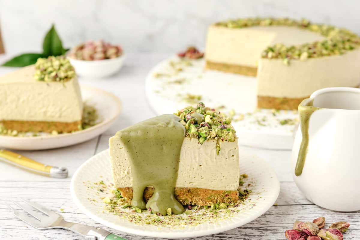 Slice of pistachio cheesecake on a plate with pistachio topping, with jar of sauce, another slice of cheesecake and rest of cut cheesecake in the background.