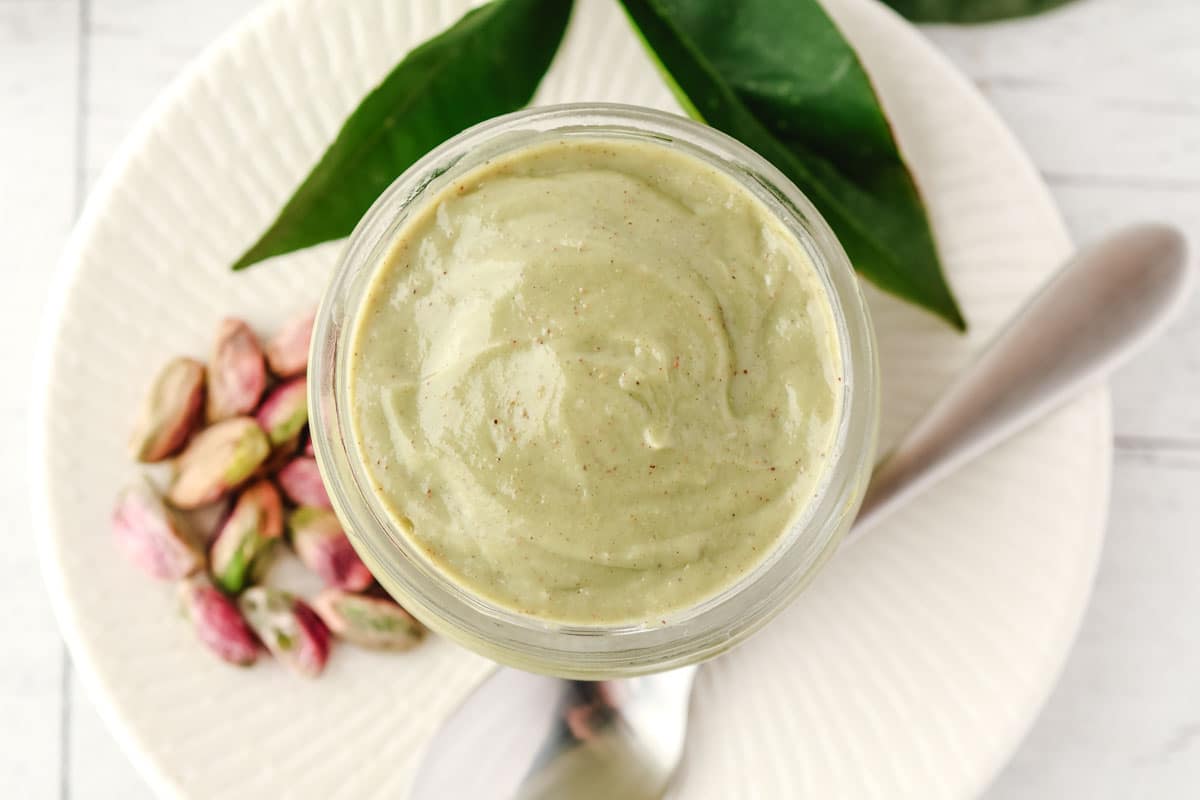 Top down shot of pistachio topping in a jar, with a spoon and pistachio nuts on the side.
