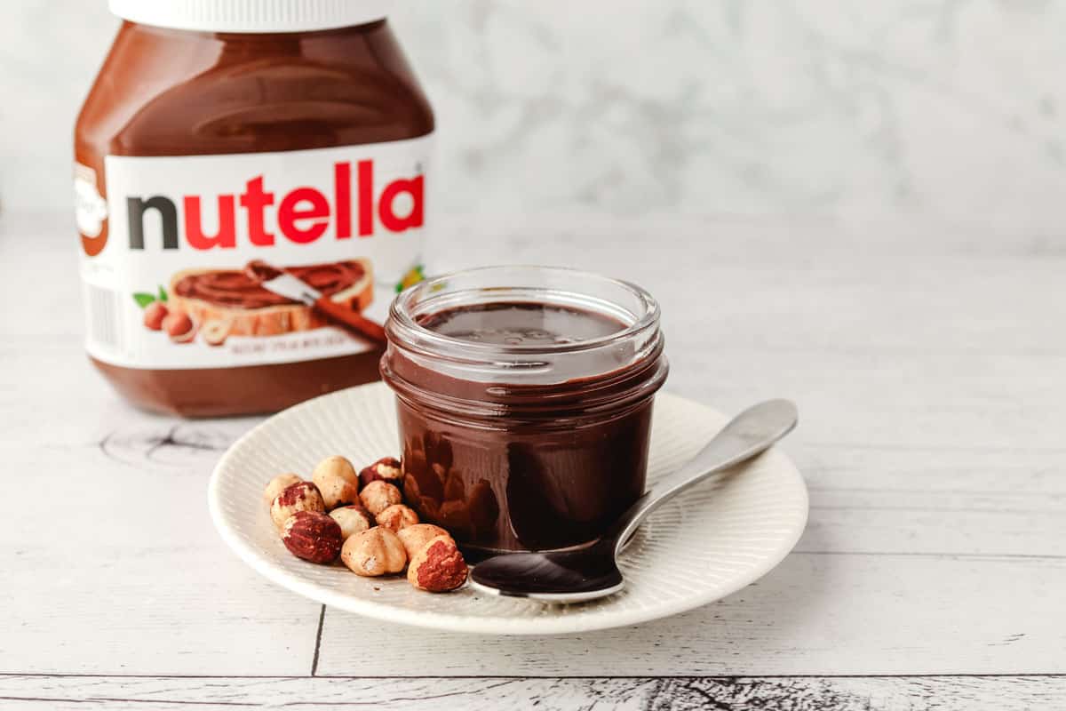 Mason jar of Nutella sauce on a plate, with a spoon and hazelnuts on the side and jar of Nutella in the background.