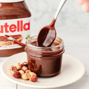 Spooning Nutella sauce out of a small mason jar, with hazelnuts on the side and jar of Nutella in the background.