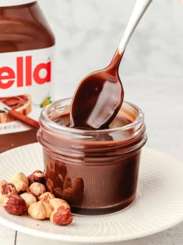 Spooning Nutella sauce out of a small mason jar, with hazelnuts on the side and jar of Nutella in the background.