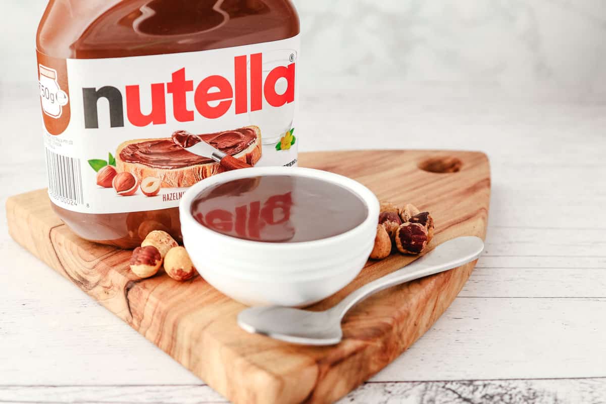 Small serving bowl of Nutella topping, with a spoon hazelnuts and jar of Nutella in the background.