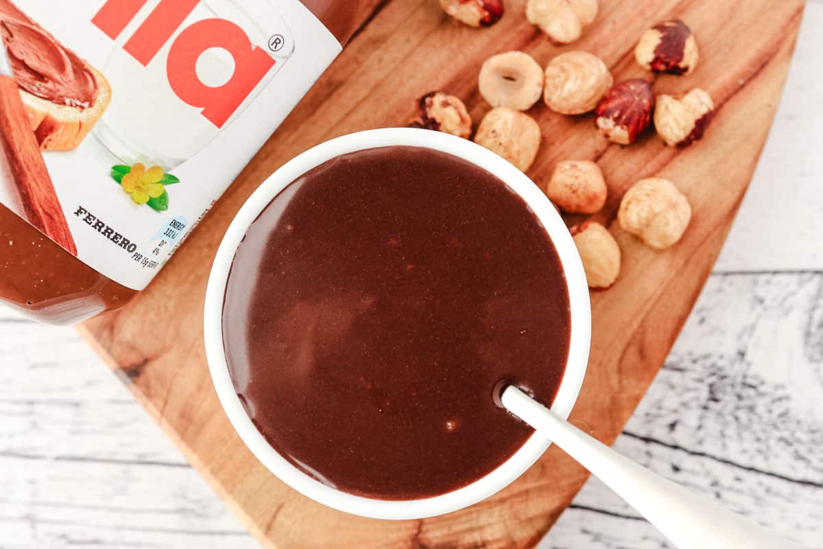 Top down view of Nutella sauce in a serving bowl with a spoon, with hazelnuts and jar of Nutella beside it.