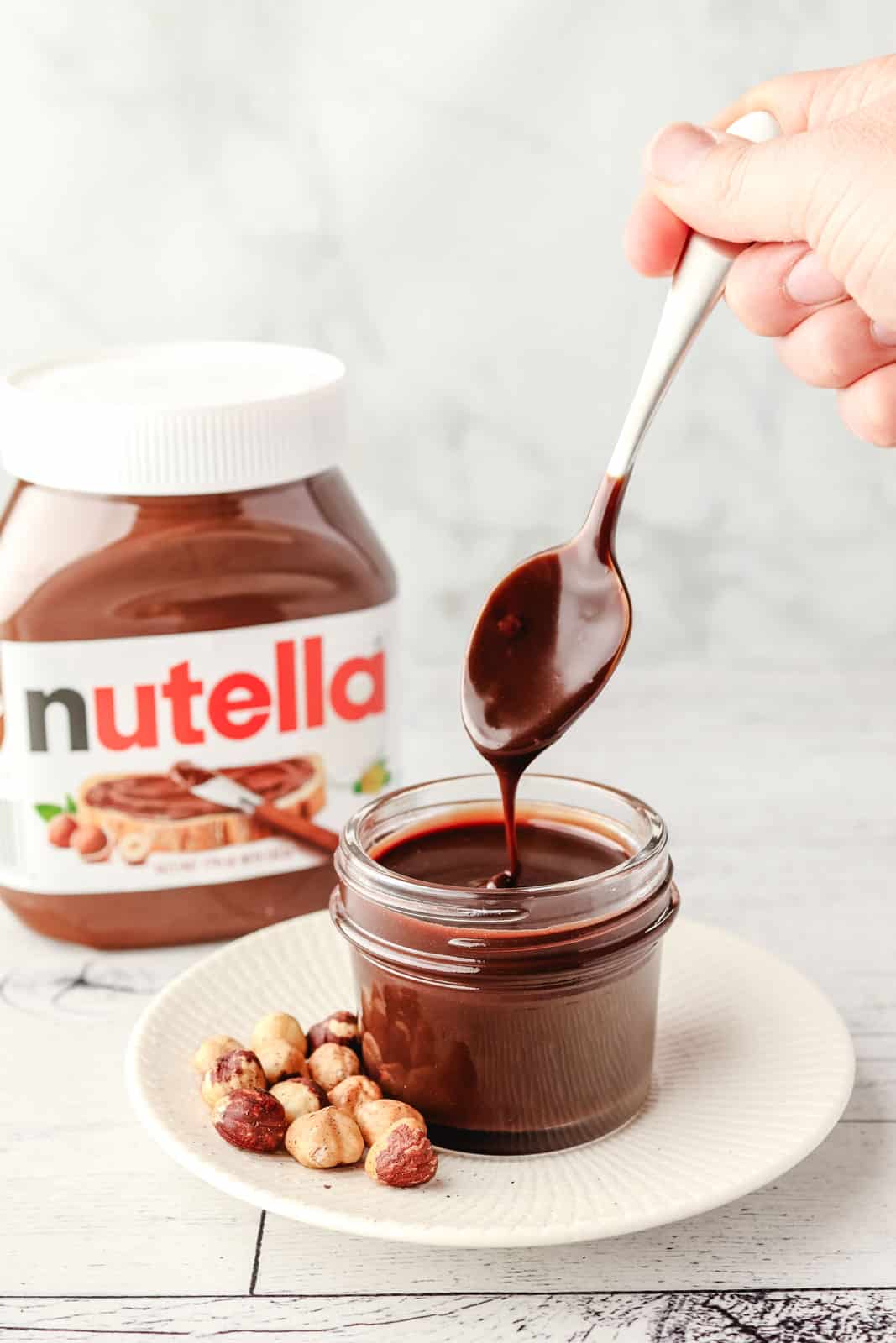 Spooning Nutella sauce out of a small mason jar, with hazelnuts on the side and jar of Nutella in the background.
