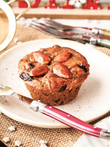 A mini Christmas cake made in a muffin tin on a plate with a fork in the side and festive decorations in the background.