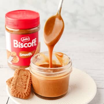 A small mason jar of Biscoff sauce with a spoon full of sauce being drizzled into it, with Biscoff cookies on the side and jar of Biscoff spread in the background.