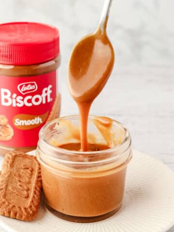 A small mason jar of Biscoff sauce with a spoon full of sauce being drizzled into it, with Biscoff cookies on the side and jar of Biscoff spread in the background.