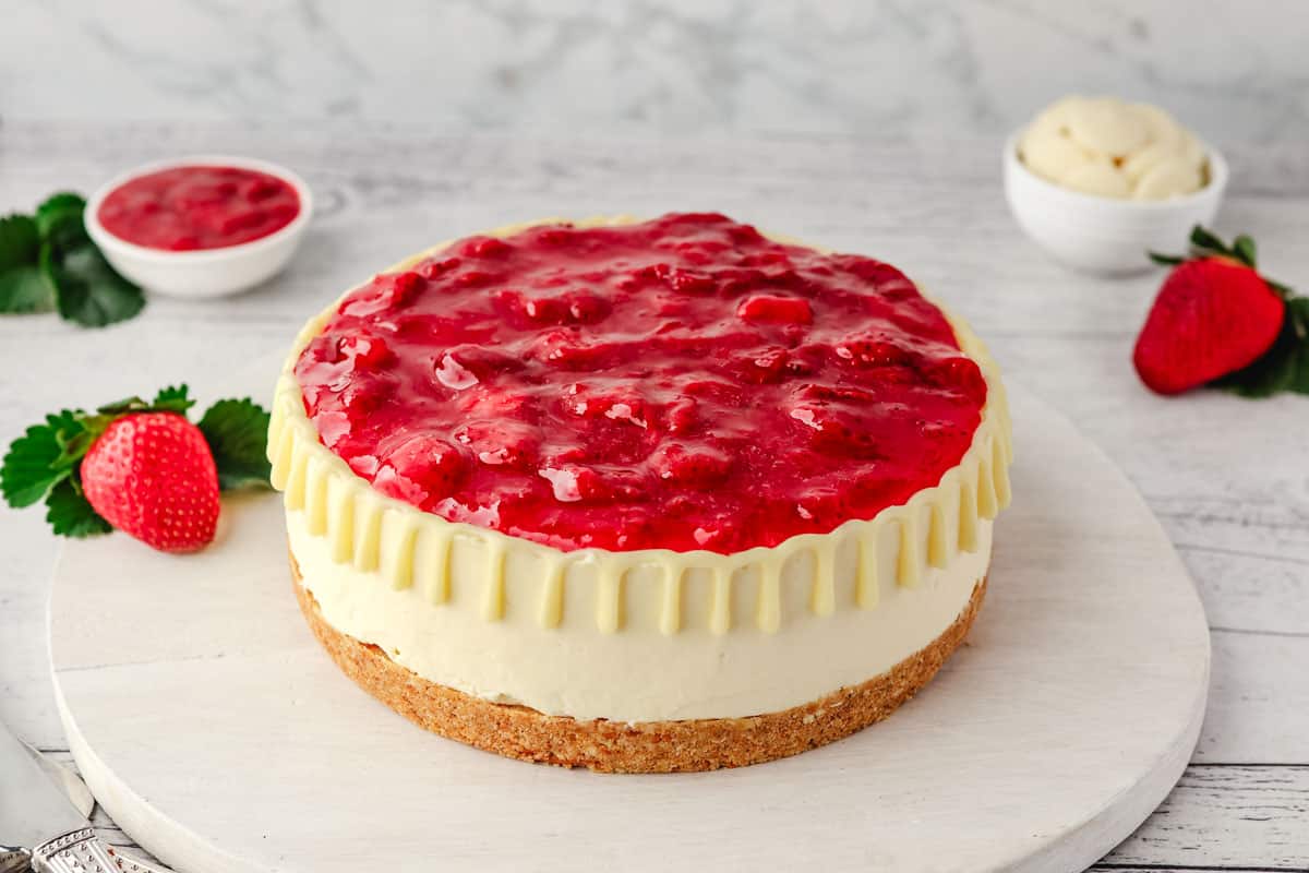 No bake White chocolate strawberry cheesecake on a serving board, with fresh strawberries, strawberry sauce and white chocolate in the background.