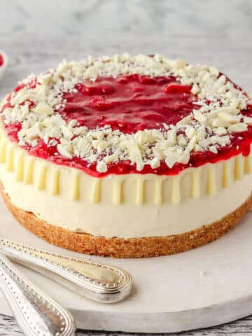 Whole strawberry white chocolate cheesecake on a serving board, with serving wear on the side.