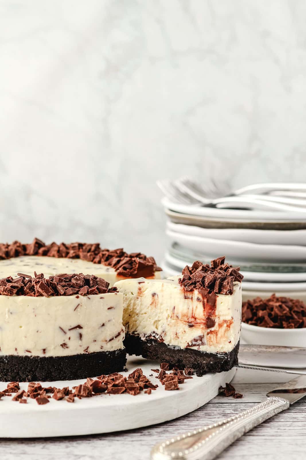 Chocolate Chip Cheesecake 2 Taking out a slice of chocolate chip cheesecake, with chocolate chips and a pile of plates in the background.